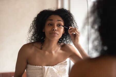 Beautiful young african American woman in towel look in mirror in bathroom paint eyelashes with black thickening mascara, biracial female get ready do makeup in home bath, beauty procedure conceptの写真素材