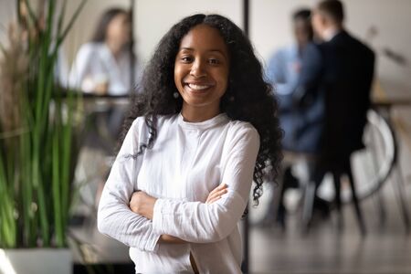Head shot portrait of smiling young african american female professional, standing with folded hands...