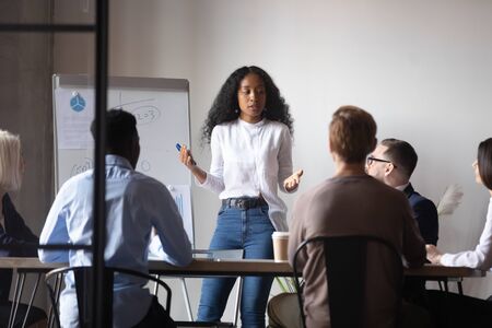 Focused african american female speaker presenting project research results to interested diverse teammates at brainstorming meeting. Confident mixed race coach giving educational lecture or workshop.の写真素材