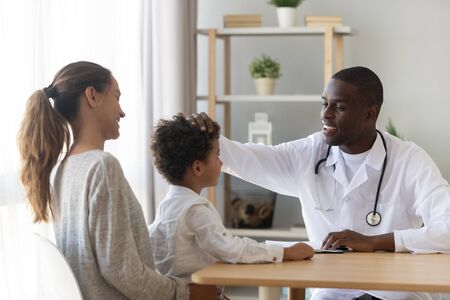 Smiling african american male doctor stroking head of little preschool cute kid patient, satisfied with medical treatment result or boys polite behavior. Happy mom with small son on medical check up.の写真素材