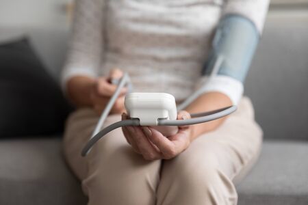 Middle-aged 50s woman sits on sofa using sphygmomanometer medical manual device measuring blood pressure at home close up view. Concept of healthcare instrument, hypertension control in aging adultsの写真素材