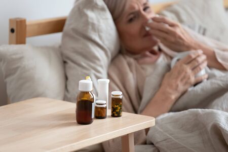 60s elderly woman lying in bed holding cup blow runny nose using handkerchief close up focus on bedside table full of antiviral medications, bottle of pills, grippe influenza, caught a cold conceptの写真素材