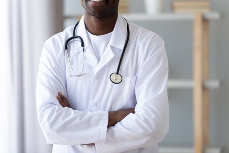 Close up cropped image young smiling professional african american male doctor in white uniform standing with folded hands and phonendoscope on neck. Modern private trustful clinic advertising.の写真素材