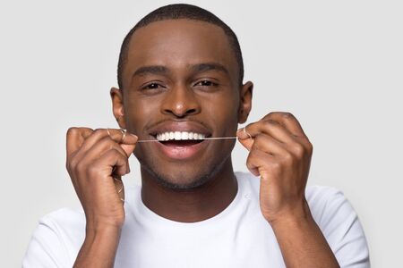 Head shot happy African American man with wide healthy smile using dental floss, cleaning teeth, looking at camera, dental care, oral hygiene concept close up, isolated on studio backgroundの写真素材