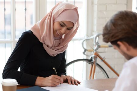 Head shot happy arabic businesswoman satisfied with negotiations results, signing agreement with business partner at office. Smiling female arabian manager putting signature on contract with client.の写真素材