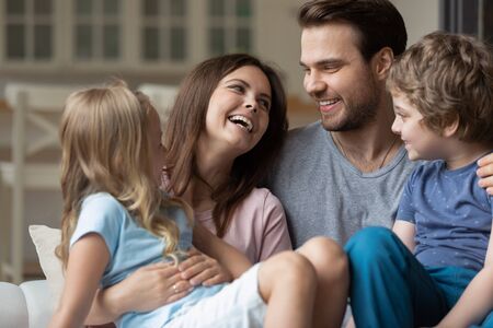 Happy married couple holding on lap little son and daughter enjoy time together on weekend free time at home, relative people laughing communicating having warm relations showing love and care conceptの写真素材