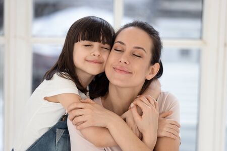 Happy young mother and little cute daughter hugging. Smiling girl with attractive mom enjoying moment together, sitting with closed eyes on couch at home. Trusted relationships between mum and girl.の写真素材