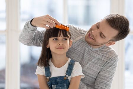Caring happy father brushing combing preschool daughter hair. Help with hairstyle in morning, smiling little girl sitting at home together playing with toy.の写真素材