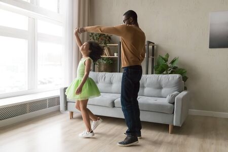 Young african American father dance together with happy preschooler daughter wearing ballet skirt, happy black dad sway swirl excited biracial girl child dancer in green dress in living room at homeの写真素材