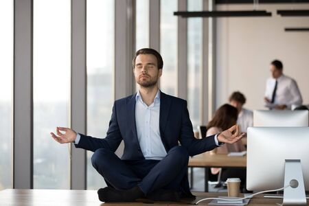 Calm businessman in suit meditating on office desk, breathing deep, enjoying break, employee practicing yoga on table at work, sitting in lotus pose at workplace in office, stress relief conceptの写真素材