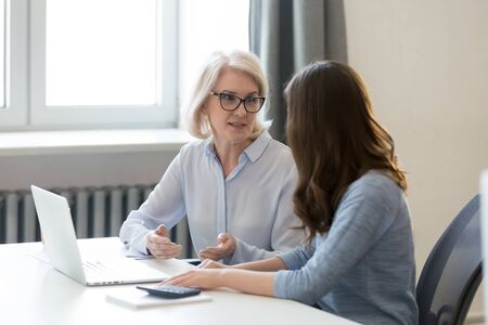 Businesswomen sit at office desk talking discussing startup project at workplace near computer, middle-aged woman speak explaining business issue to colleague or intern, working together at laptopの写真素材