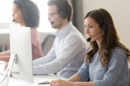 Millennial female call center agent wear headset with microphone consult customer online on pc in coworking space, focused woman employee in earphones busy working on computer in shared officeの写真素材