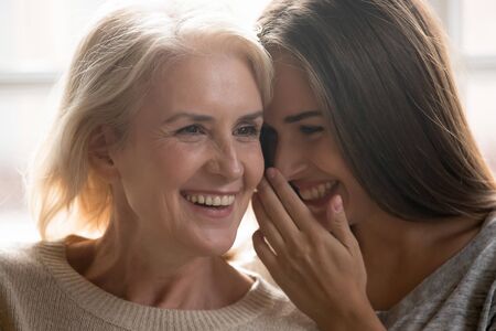 Close up of smiling millennial female whisper tell secret in senior mother ear, happy senior mom and adult daughter have fun talking gossiping at home, excited two generations of women share thoughtsの写真素材