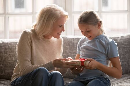 Loving middle-aged 60 grandmother sit on couch greeting congratulate small granddaughter with...