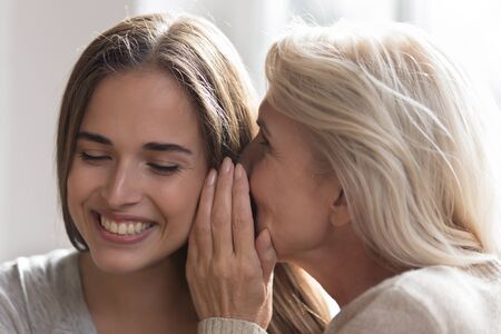 Loving elderly female senior mother whisper in smiling adult daughter ear sharing secret, tell hidden information, happy mom and child two generations of women gossip chat at home laughingの写真素材