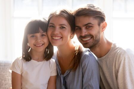 Head shot portrait of smiling family, happy mother, father and adorable little daughter sitting together, cuddling, posing for photo, looking at camera, enjoying free time, weekend at homeの写真素材