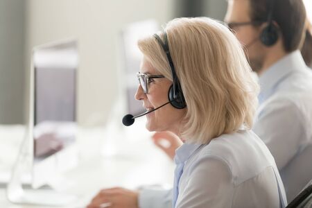 Close up of middle-aged woman call center agent wearing headset speak consult client online at helpdesk, senior female operator work at helpline in customer service shared office using computerの写真素材