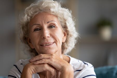 70s grandmother seated indoors posing folded arms under chin looking at camera, representative of baby boomer generation, older people retired well-being life, kindly loving granny close up portraitの写真素材