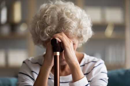 Close up view sad 60s woman bowed her head hiding face with hands holding walking cane stick, aggrieved about illness physical damage, chronic progressive movement disorder Parkinson disease conceptの写真素材