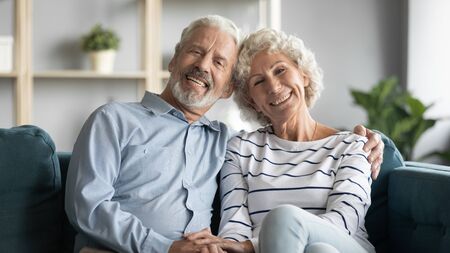60s loving couple sitting in living room posing looking at camera smiling capture moment for family...