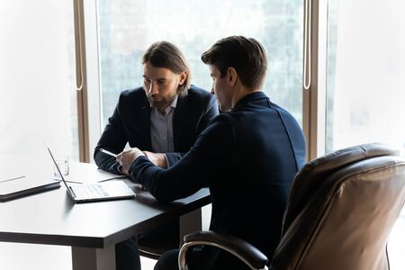 Concentrated businessmen sit at office desk look at laptop screen discuss business project together,...