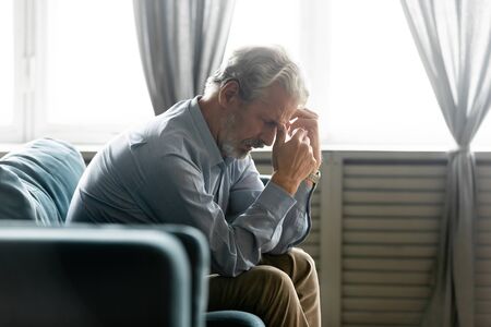 Side view in living room seated on couch hunched old 50s male hold hands near face looking desperate and lonely feels worried. Concept of illness and diagnosis, older generation humans life troublesの写真素材