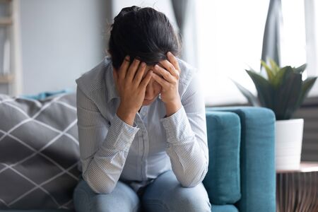 Depressed frustrated young indian woman hide face in hands suffer from mental stress, life problems or headache, upset worried ethnic teen girl think of grief feel desperate sit alone on sofa at homeの写真素材