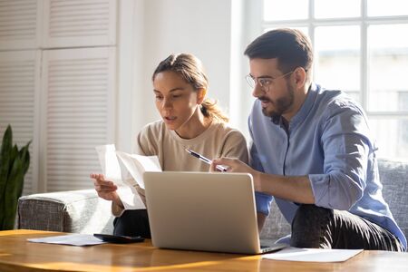 Concentrated young couple sit on couch busy managing finances paying bills on computer online, focused anxious millennial husband and wife calculate household expenses or taxes use internet bankingの写真素材