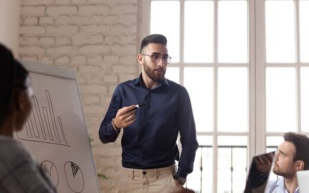 Young Arabian male speaker or coach talk with diverse employees explain make flip chart presentation at briefing, millennial Arabic businessman speak with workers present plan on whiteboard at meetingの写真素材