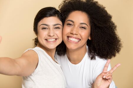 Camera view head shot overjoyed indian happy millennial girl taking selfie with smiling african american young female friend, demonstrating positive emotions isolated on yellow studio background.の写真素材