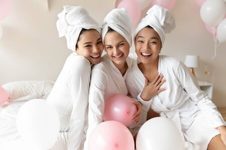 Portrait of overjoyed multiracial female friends in bathrobes and towels have fun enjoy bachelorette bridal shower in bedroom, smiling multiracial millennial girls pose celebrate hen party togetherの写真素材