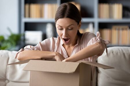 Overjoyed millennial girl sit on sofa in living room unpack cardboard parcel with online Internet order, happy young woman client customer open carton box shopping on Web, good delivery conceptの写真素材