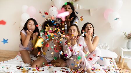 Happy multiracial millennial girls in pajamas have fun in bedroom blow colorful glitter confetti...