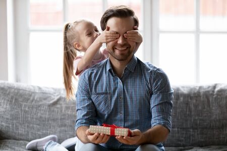 Excited little girl close eyes congratulate overjoyed young father present gift box at home, happy small daughter make birthday surprise for smiling dad give wrapped beautiful package greetingの写真素材