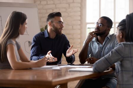 Thoughtful millennial multiracial colleagues sit gather at office desk discuss business ideas at...