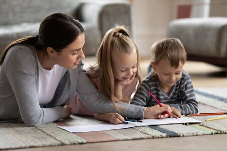 Loving mom or nanny lying on warm floor in living room with cute little kids drawing together, caring mother have fun enjoy time with small children painting picture, family weekend conceptの写真素材