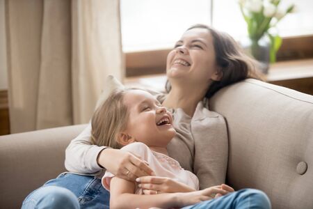 Happy mother and laughing little daughter relaxing on couch, young mum and adorable preschool child girl spending free time together at home, having fun, embracing, lying on cozy sofa close upの写真素材