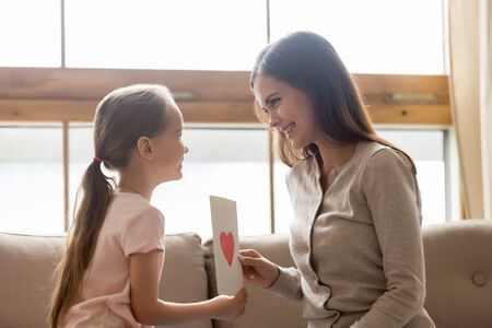 Smiling mother receiving gift from little adorable daughter at home, cute girl congratulating mum...