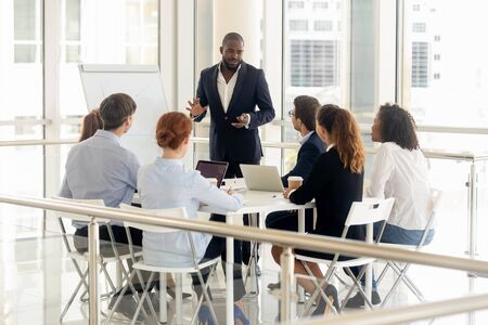 Smiling african American businessman stand make flip chart presentation for diverse work team in office, black male coach or trainer talk present strategy on whiteboard, training employees at meetingの写真素材