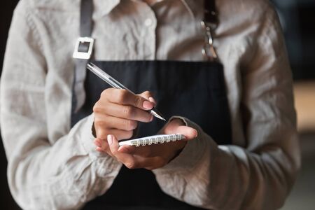 Close up of waitress in apron hold notebook taking order from clients in cafe or restaurant, female staff wearing uniform writing down serving customer in coffeehouse. Good service conceptの写真素材