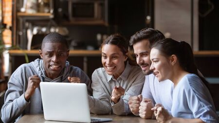 Excited diverse millennial friends gather in bar watching football match on laptop online, overjoyed hopeful fans young people hang out have fun in cafe cheering favorite sports team for win in gameの写真素材