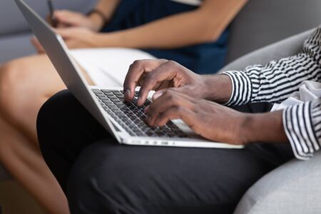 Close up of african American man sit on couch in office texting on laptop, mixed race ethnicity biracial male busy typing browsing internet in computer, employee surfing wireless web using gadgetの写真素材