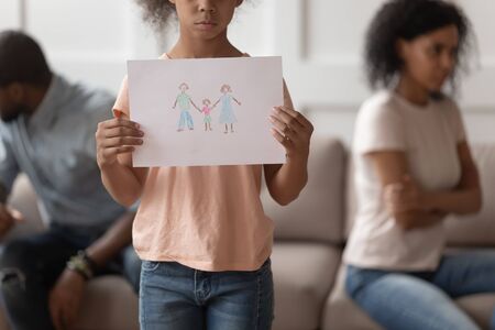 Close up cropped image african american girl holding hand drawn picture with happy family, blurred separate parents on background, family conflicts, break up, divorce concept.の写真素材