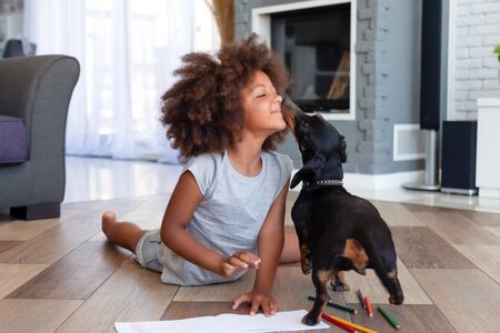 Funny little African American girl lying on floor coloring picture having fun with dog, family pet kissing playing with small child painting at home, kid laugh entertaining with domestic animalの写真素材