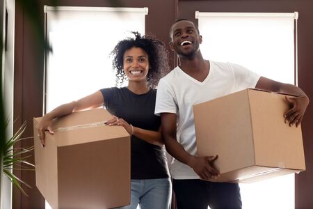 Overjoyed black man and woman carrying carton boxes with personal belongings impressed entering own...
