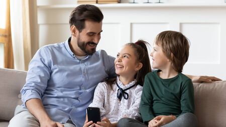Smiling young Caucasian dad sit on couch talk laugh with little kids use smartphone together, happy...