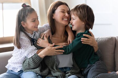 Happy little kids sit on couch hug cuddle excited young mom or nanny show love and care, overjoyed small children relax with mother embrace share sweet tender family moment in living room at homeの写真素材