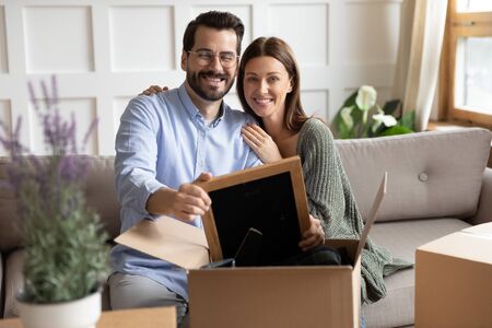 Portrait of smiling young husband and wife sit on sofa in living room unpack cardboard boxes with...