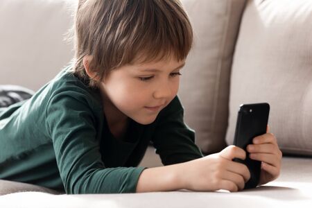 Cute smart little boy lying on couch in living room playing game on modern cellphone gadget, small...