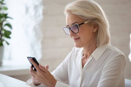 Focused attractive middle-aged businesswoman in glasses seated at workplace desk holding smart phone read latest news, check e-mail, using business application, typing message solve issues distantlyの写真素材